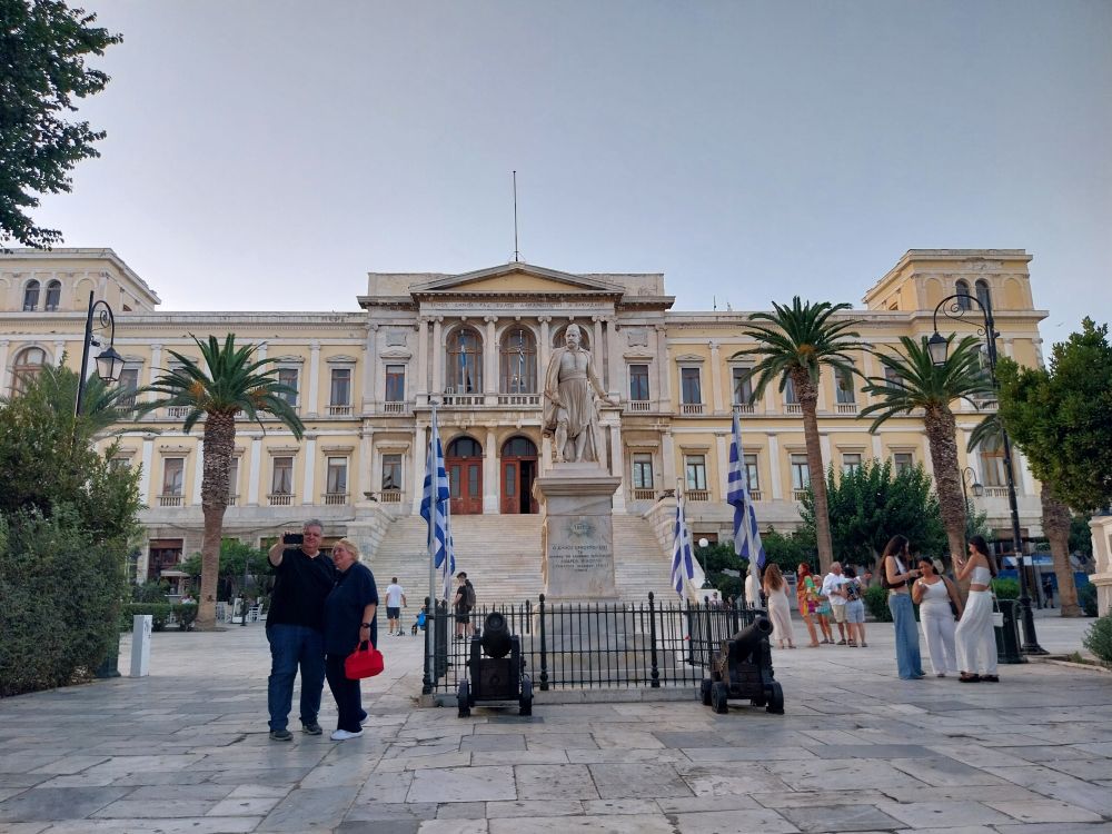A grand building in the neoclassical style in a cobbled square filled with people hanging out. Palm trees are lining the square. 