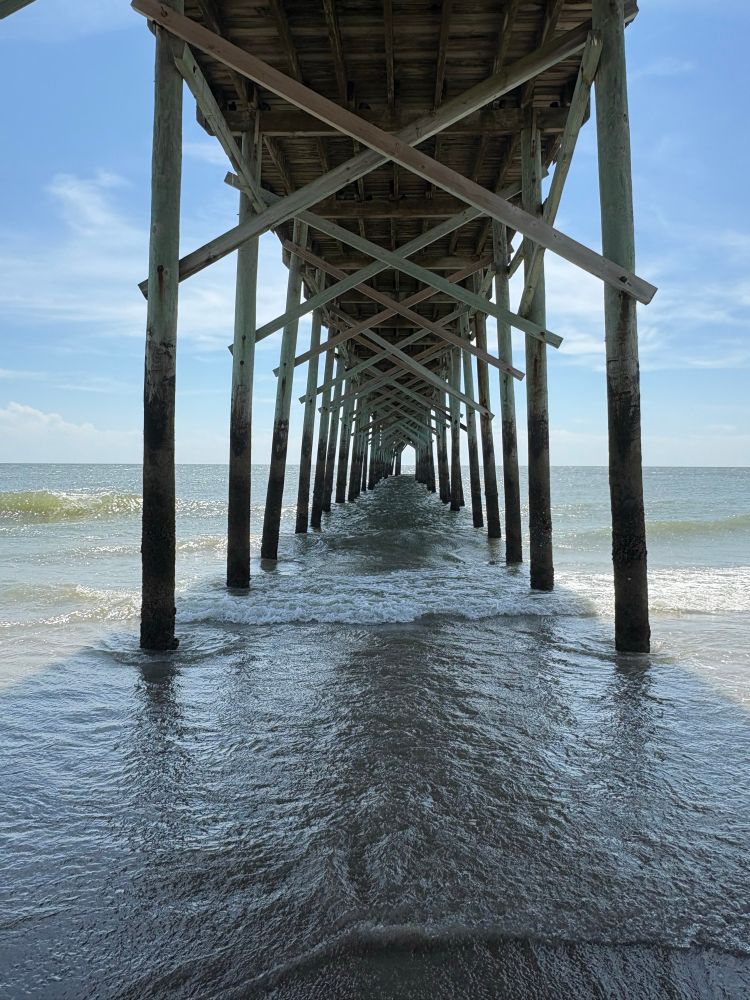 Pier pillars make an abstract tunnel out to sea, with shallow waves across the sand below. 