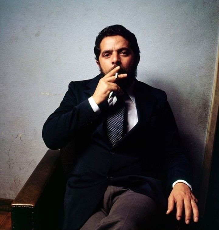 Foto do presidente Lula jovem,.de terno, fumando um cachorro garro. Um homem de barba e cabelos pretos terno preto e gravata cinza