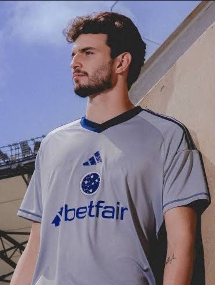 Foto de um homem branco de barba olhando ao Léo com o terceiro uniforme de goleiro do cruzeiro, que é em tom acinzentado com escudo e patrocínio azul escuro, se assemelhando ao uniforme do CEFET MG