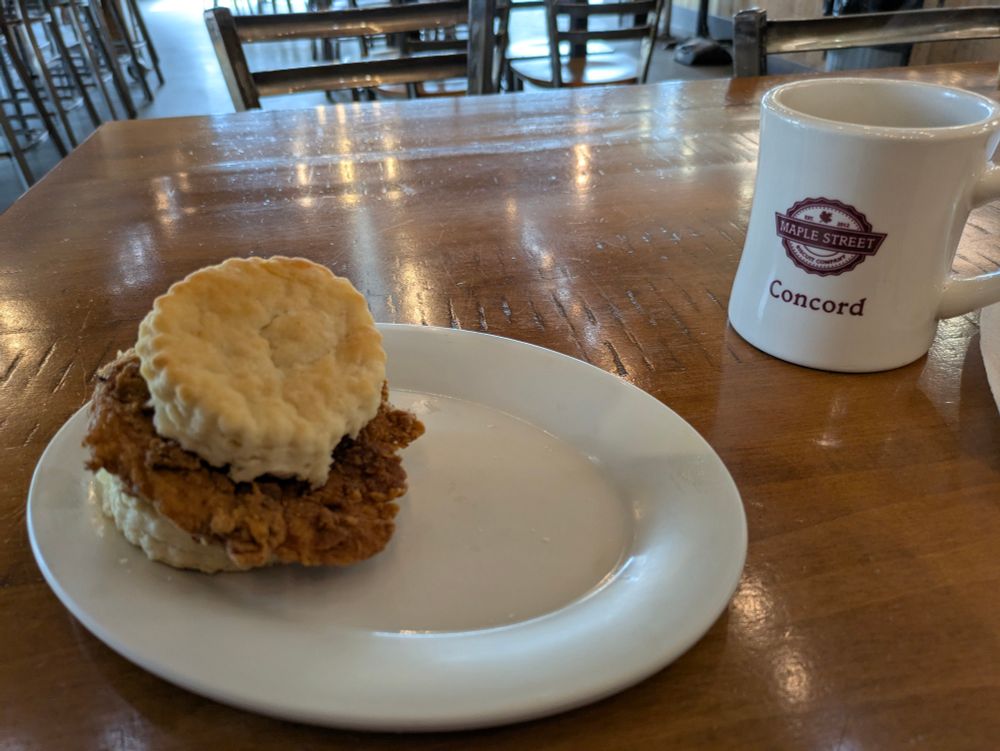 A piece of fried chicken on a biscuit along with a cup of coffee 