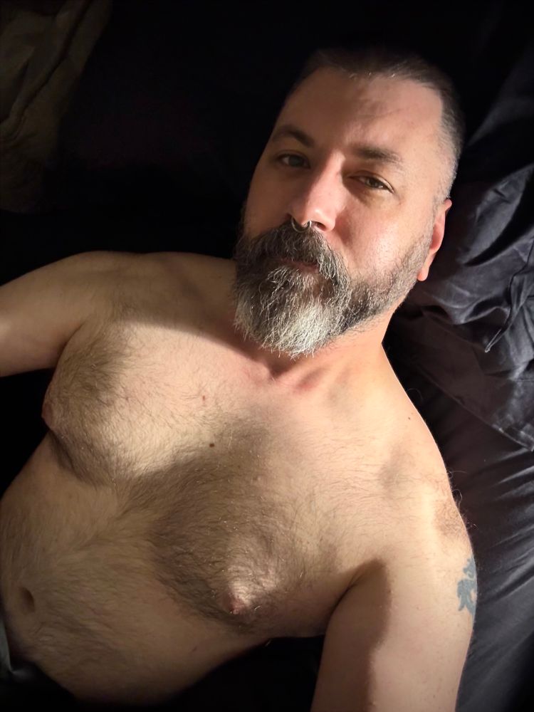Shirtless salt & pepper bearded man laying on a dark bed spread. 