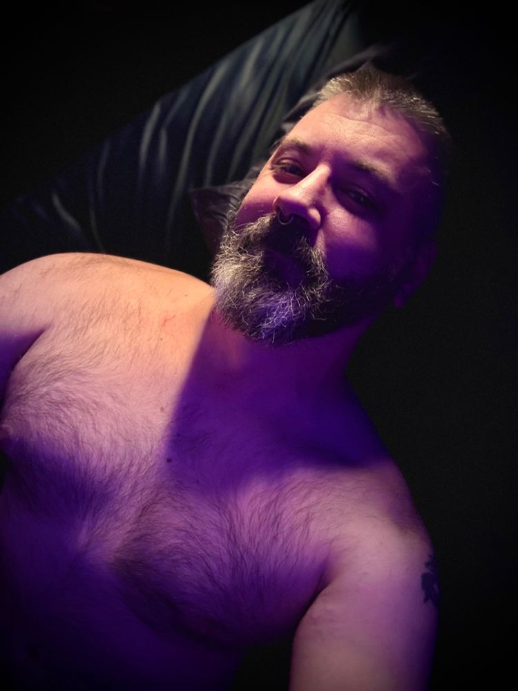 Shirtless bearded man lying in dark sheets in bed smiling up tiredly at the camera. There is warm light streaming from above and purple light in the shadows. 