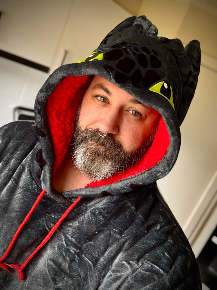 Bearded smiling man with salt/pepper wearing a Toothless oodie from How to Train Your Dragon. 