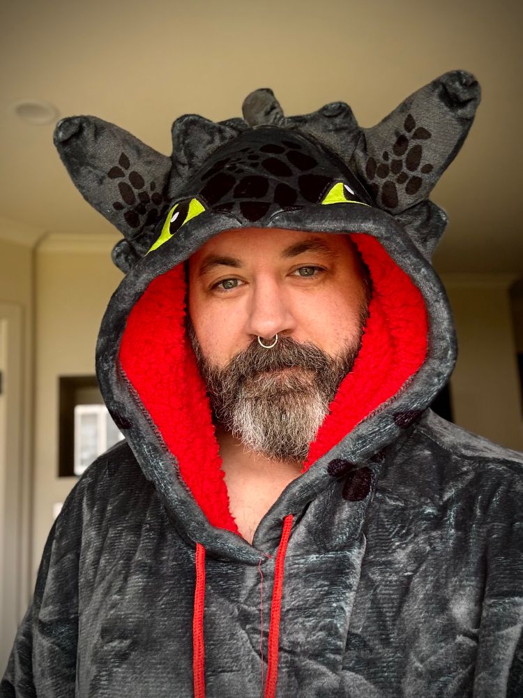 Bearded smiling man with salt/pepper wearing a Toothless oodie from How to Train Your Dragon. 