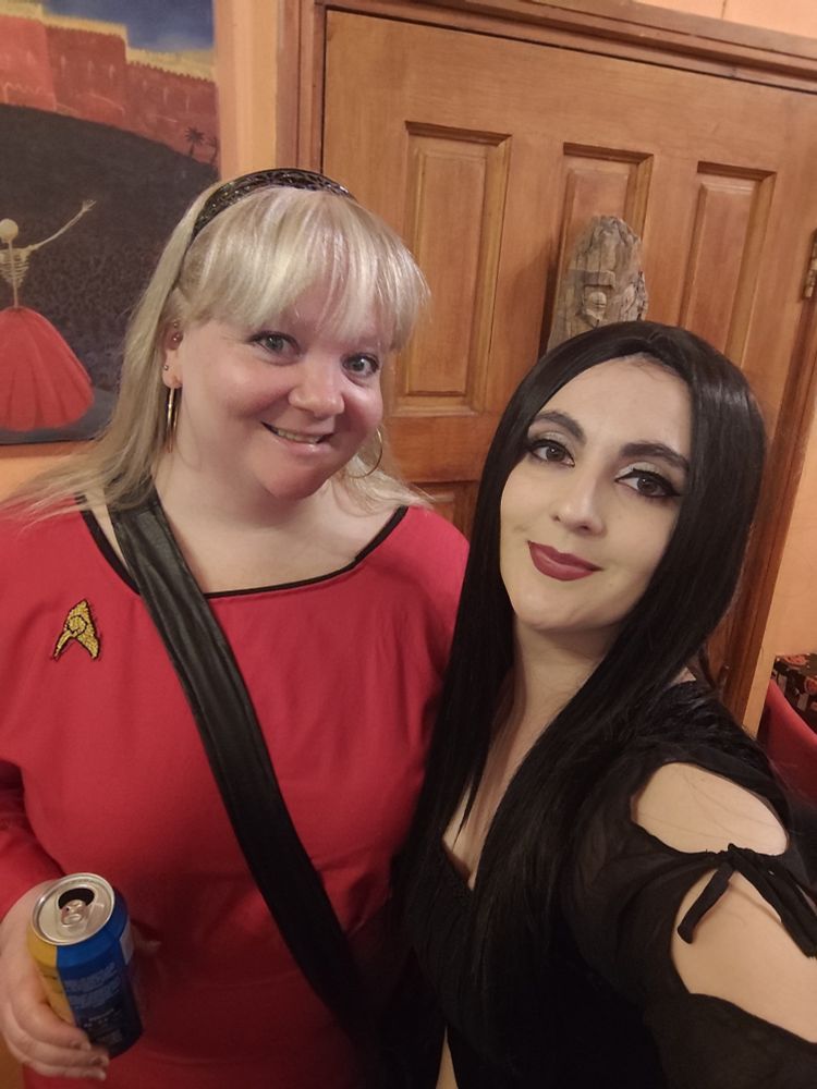 Photo of two women smiling at the camera. The one on the left (me) is wearing a homemade Star Trek Original Series uniform and holding a can of beer, and the one on the right is dressed as Morticia Addams and talking the photo.