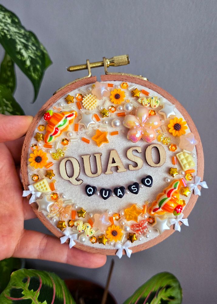 A hand holding a bead embroidered hoop. The wooden letters read "quaso". The letter beads underneath also read "quaso".