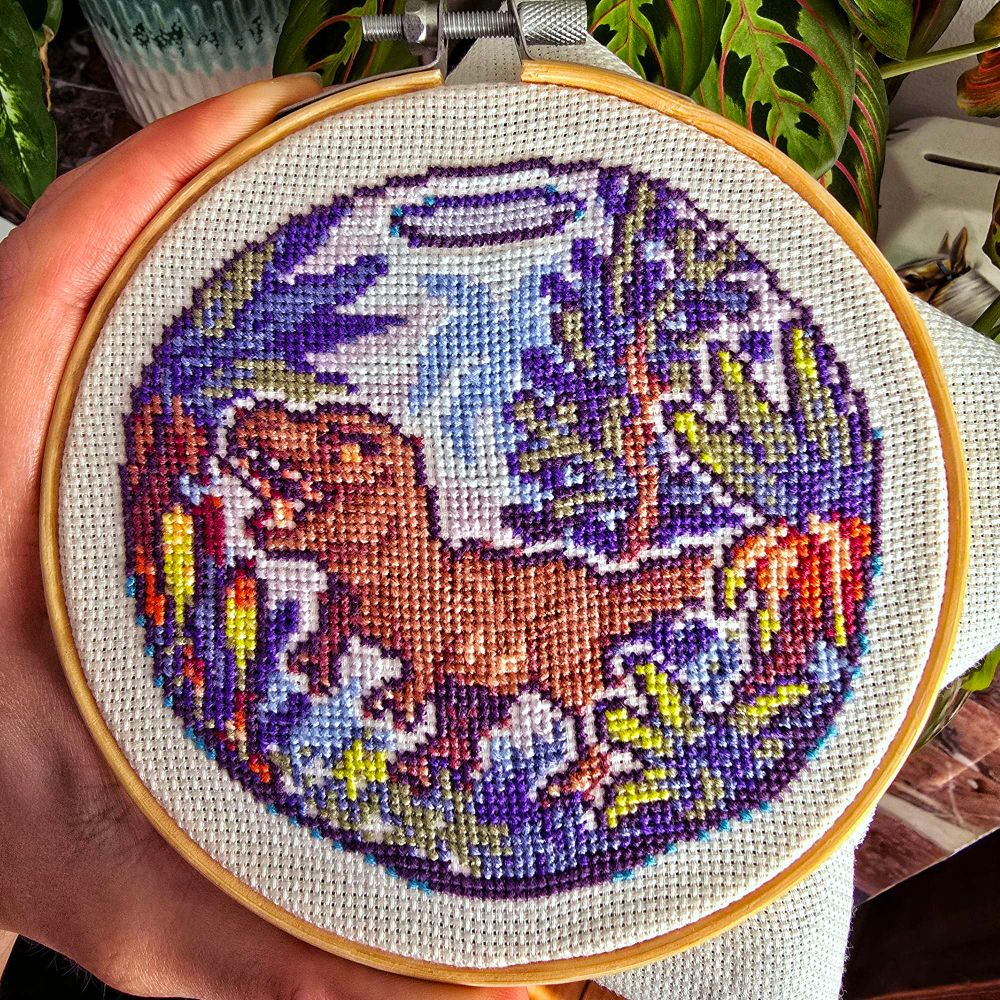 A hand holding an embroidery hoop with a finished cross-stitch.

The cross-stitch depicts a tiny T-Rex in a terrarium, surrounded by various plants and trees.