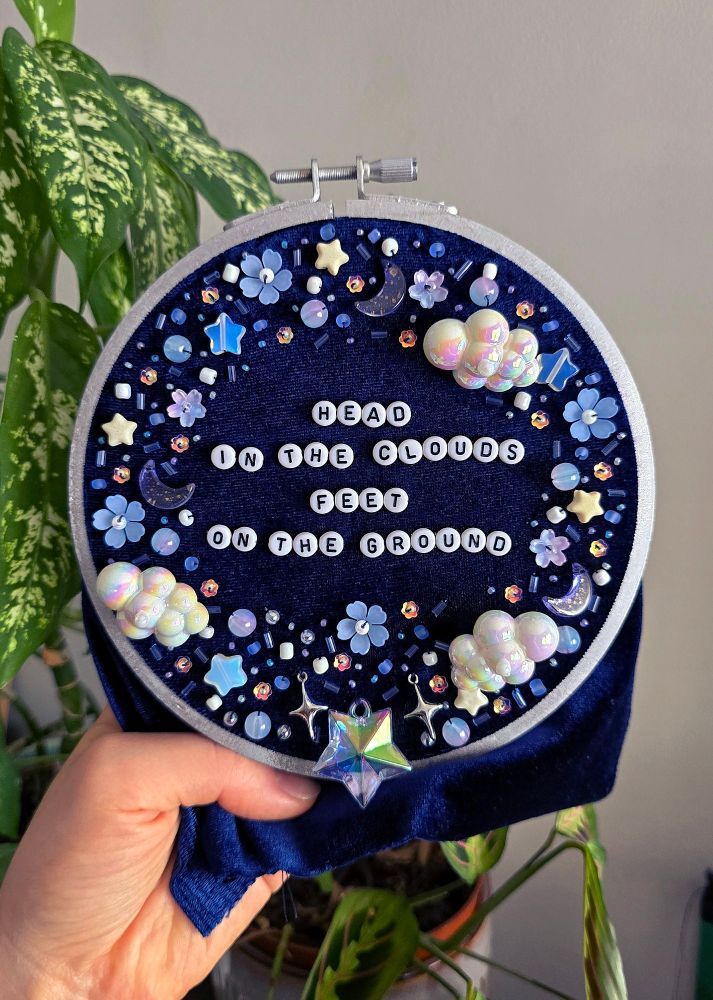 A hand holding up a silver-painted embroidery frame with bead embroidery on dark blue velvet. The text reads "head in the clouds, feet on the ground".
