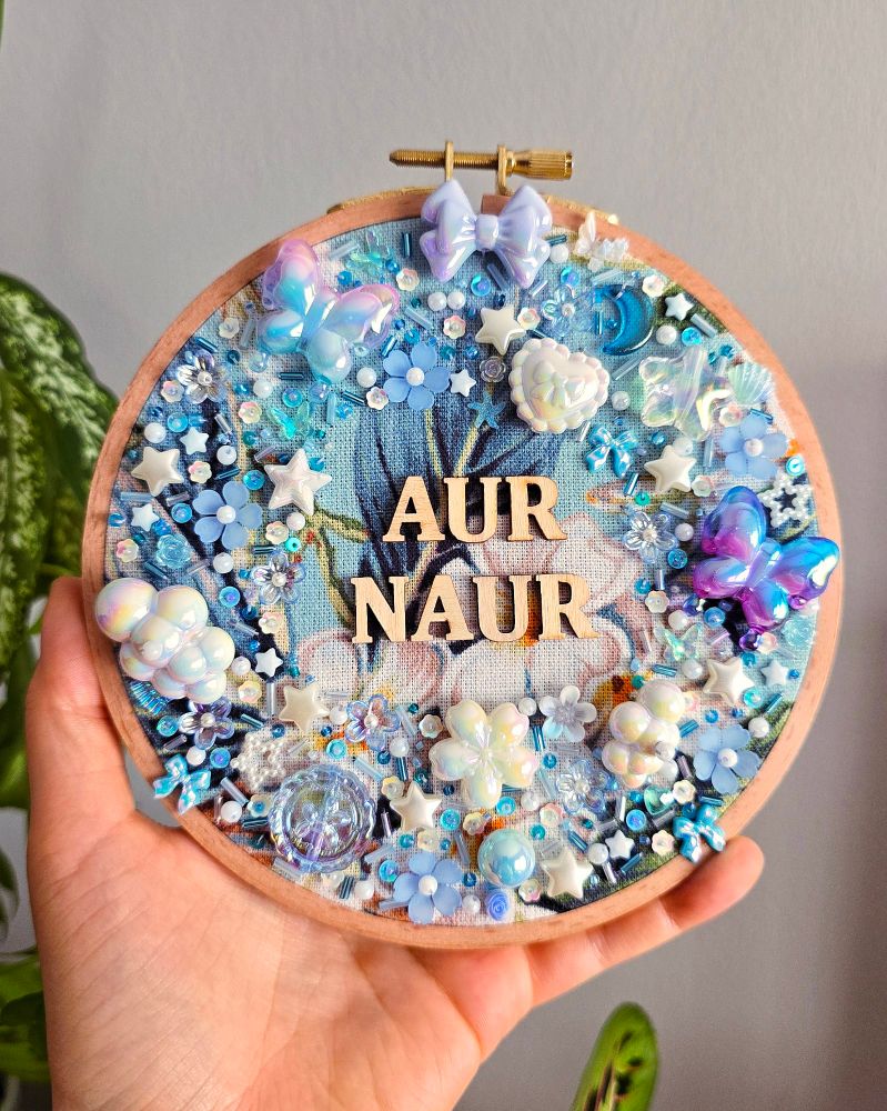 A hand holding a bead embroidered hoop. The wooden letters read "aur naur".