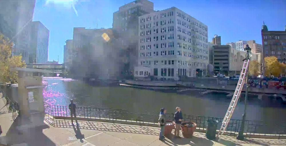 A view from the Bronze Fonz cam showing the downtown Milwaukee Riverwalk, river, and buildings. On the left in this segment of Riverwalk is the Bronze Fonz statue. At the right are two men looking up at a lamppost that has a ladder leaning against it.