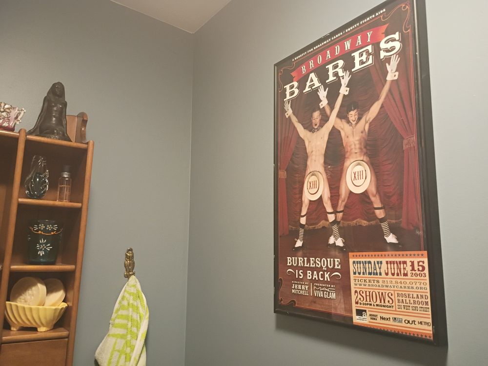 A bathroom with a "Broadway Bares" poster with some dudes doing burlesque.