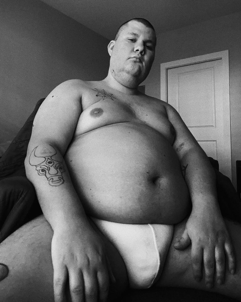 B sits reclined in a chair in a white jockstrap. 