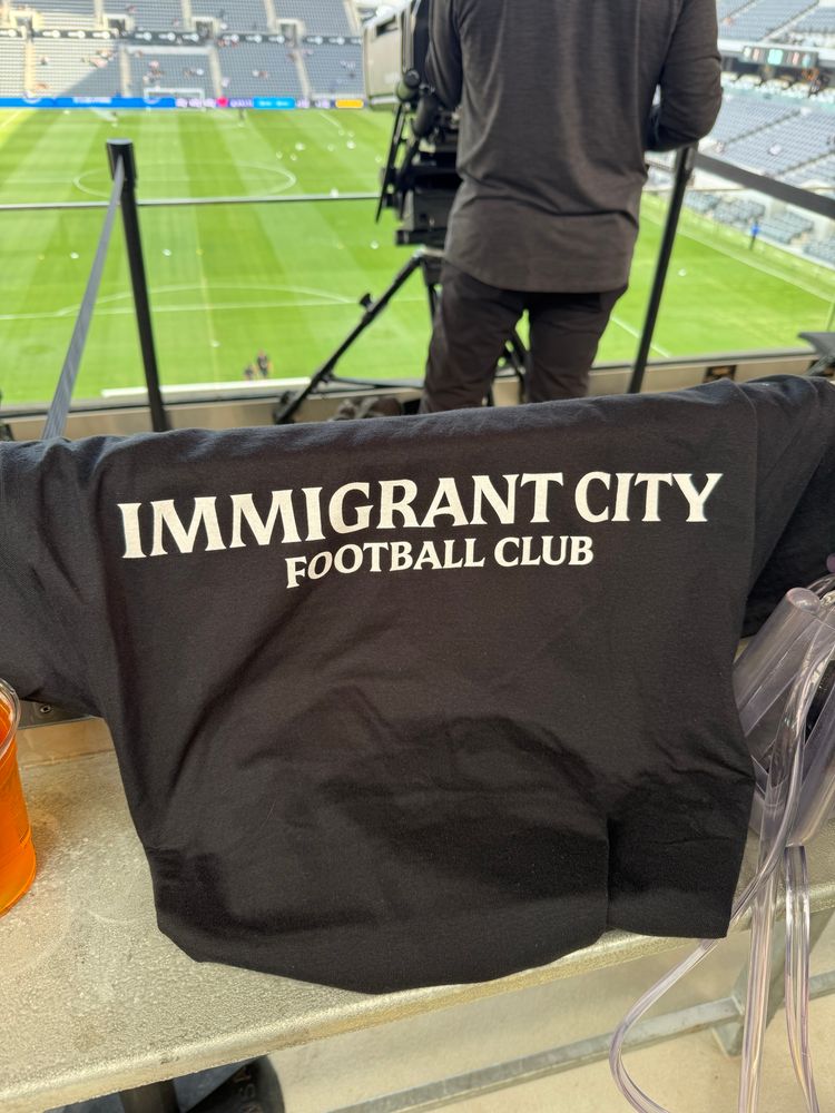 front of black shirt in white font that says IMMIGRANT CITY FOOTBALL CLUB