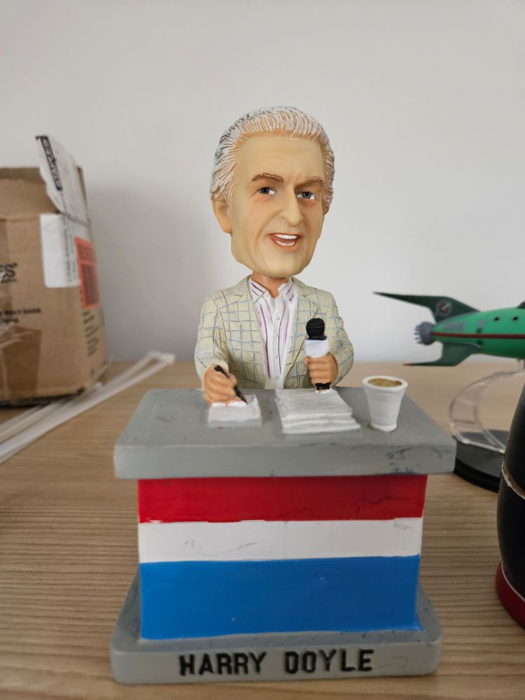 A photo of a bobblehead of a man with white hair, in a blazer with a microphone and a pen, labeled "Harry Doyle" 