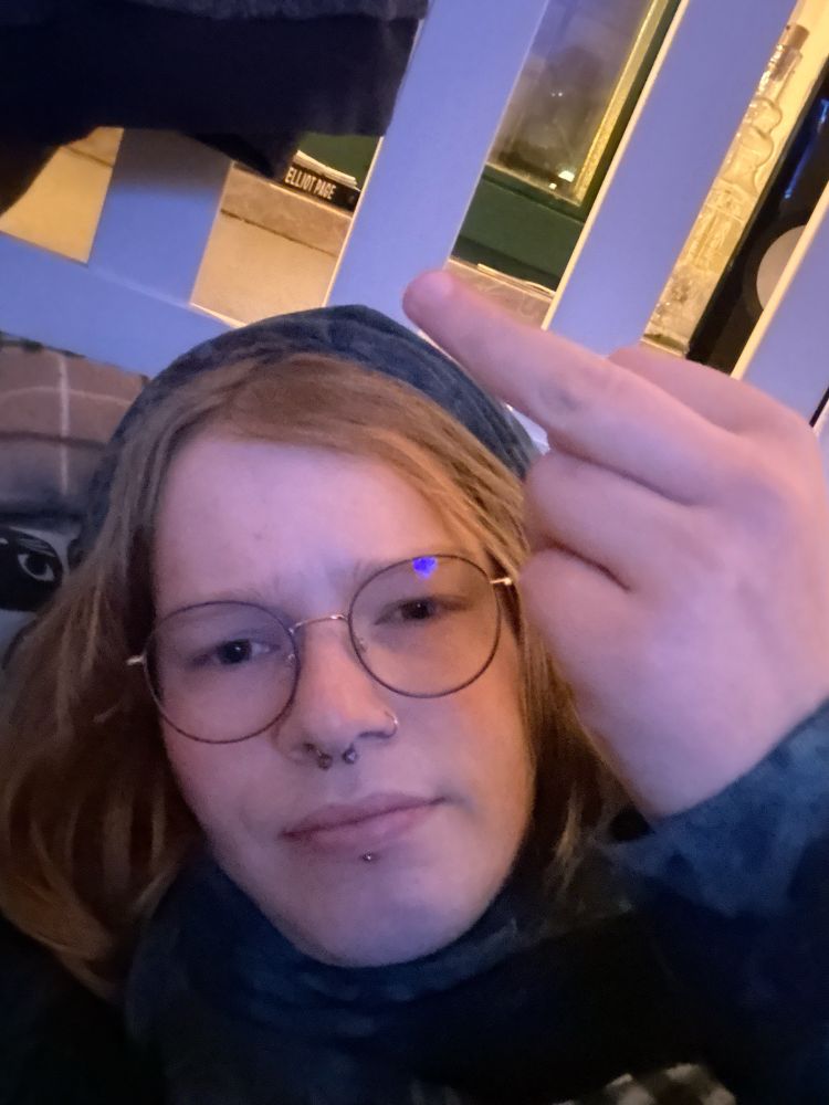 Fay liegt im Bett und schaut den Mittelfinger zeigend in die Kamera. Sie*er hat die Kaputze eines Hoodies auf.