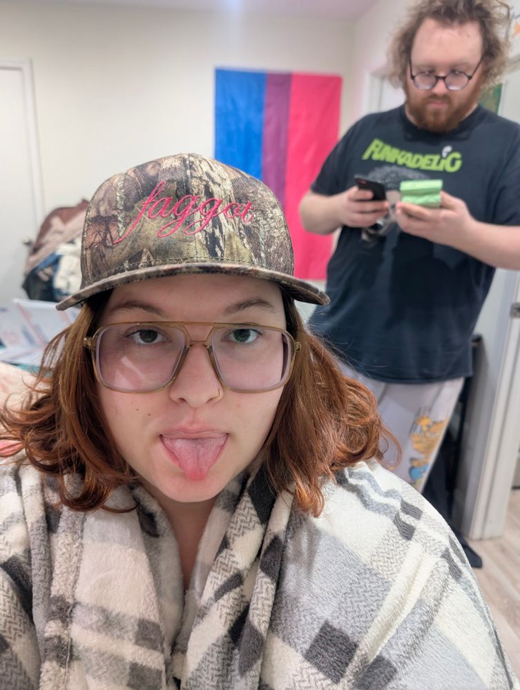 White person with long orange hair and brown glasses wears a mossy oak camo hat with "faggot" printed in bright pink script across it.  Their boyfriend stands in the background on his phone.