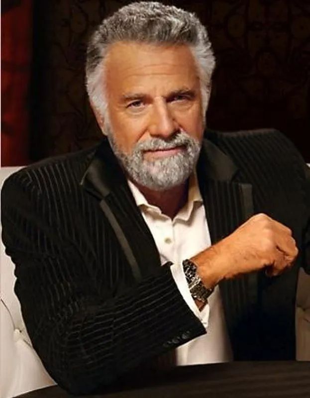 Still shot of the "most interesting man in the world" from the Dos Equis commercial. He is an older man, with grey hair and beard, sitting at a table with a black jacket over white dress shirt.