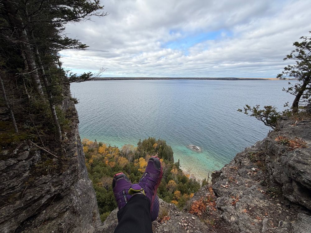 A pair of feet in purple hiking shoes rests on a rocky ledge high above a calm lake. The view includes trees with autumn foliage and a cloudy sky, with clear blue water transitioning to shades of green near the shore.