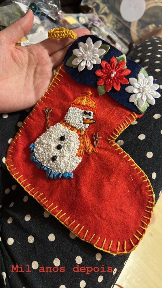 Foto de uma bota vermelha de feltro costurada em ombro caseado em fio amarelo ouro e com um bordado de boneco de neve e detalhes de flores na parte de cima.