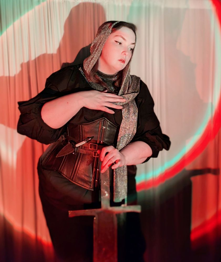 A plus sized pale woman taking a Joan of arc inspired ootd. She has a silver metallic hood and collar over a black blouse, leggings and leather corset. She has a wee knife and big sword for the vibes 
