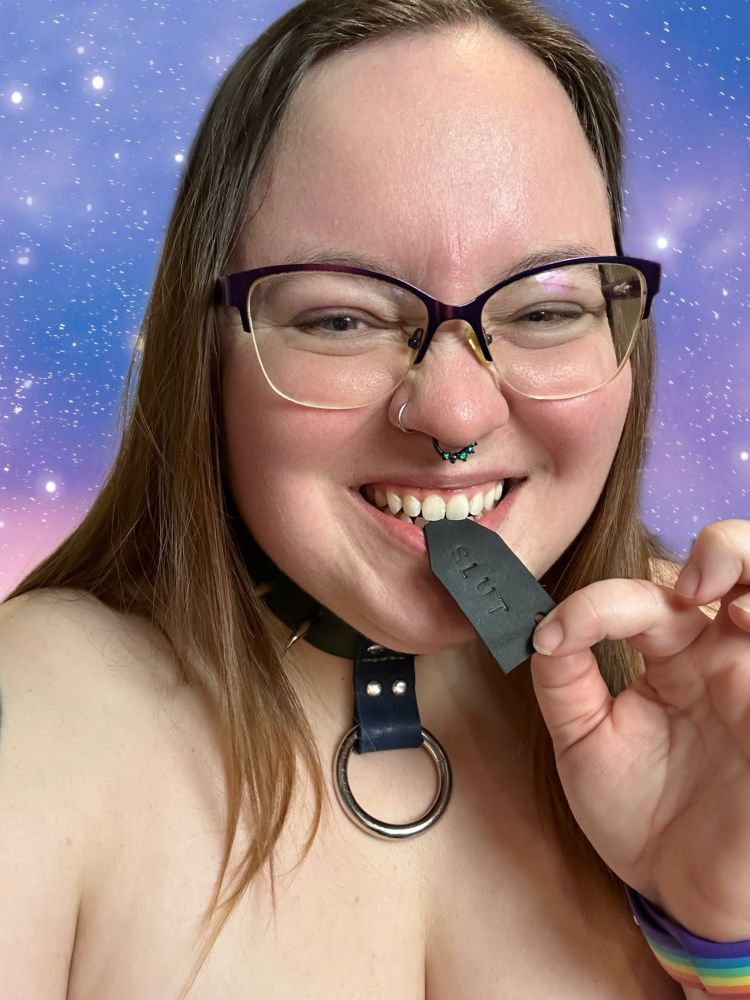 Leth showing off their new collar made by DadeAudio. It’s green with a blue centerpiece connected to an O ring, and two claw shaped pieces on each side of the centerpiece. They’re holding a small piece of black leather with the word “slut” stamped into it between their teeth