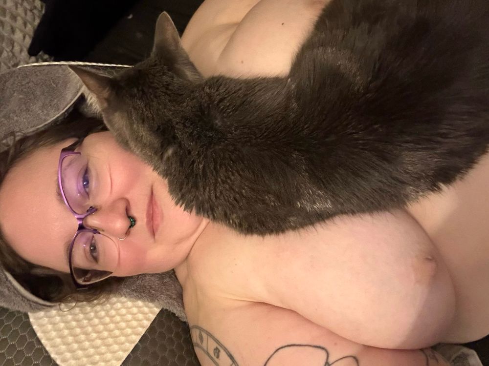 Leth laying down topless with their cat, Tina, laying between their tits and using Leth’s cheek as a pillow
