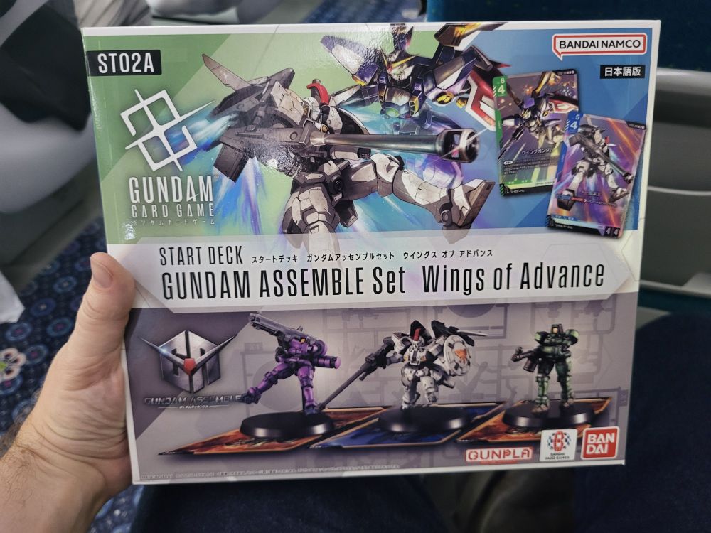 My hand holding the starter set for the Gundam Card Game including the Gundam Assemble miniatures set with the Talgeese and two Leos.