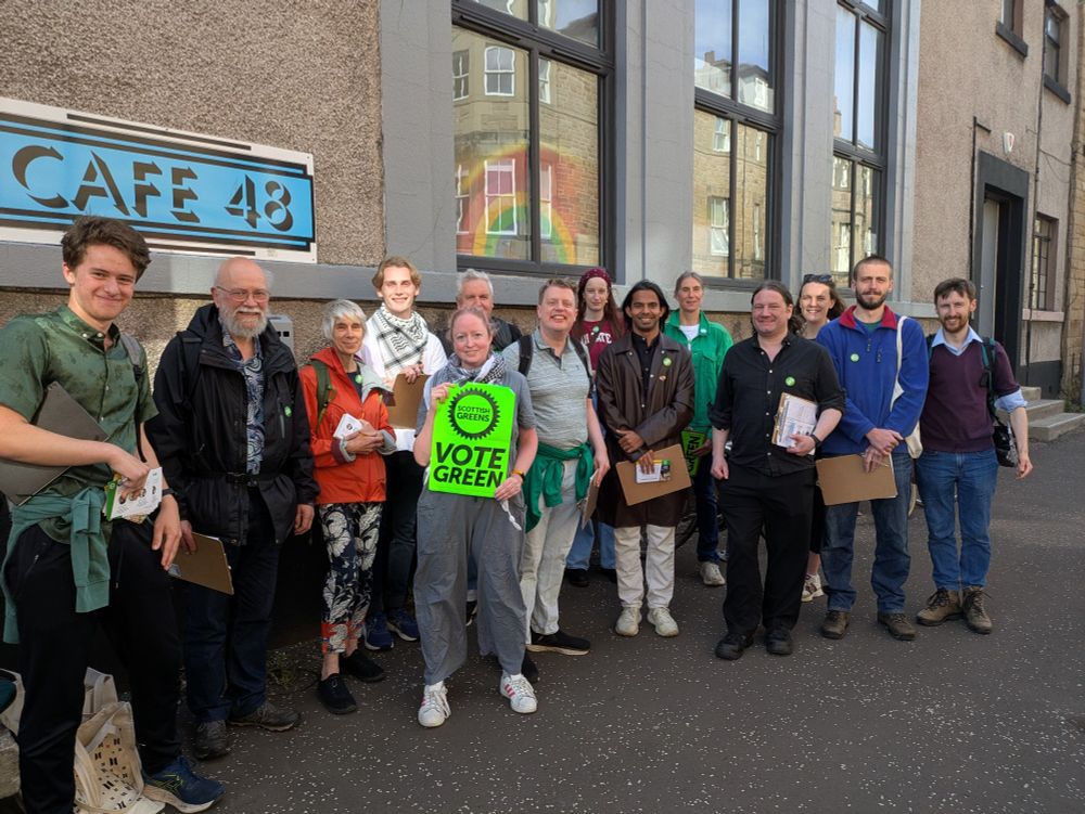 14 smiling green activists surround candidate Q in the middle, standing outside a building. Kate is holding a bright green vote green poster