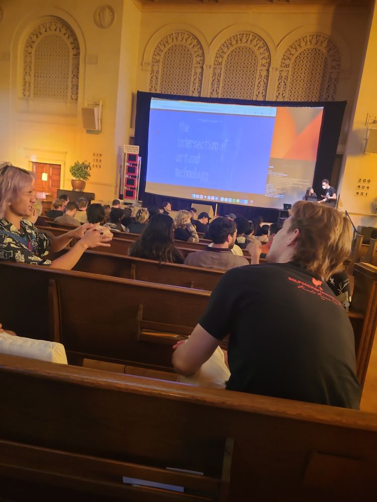 Bros discuss art and technology in an auditorium. A projector screen says "the intersection of art and technology" in very low contrast ascii art.