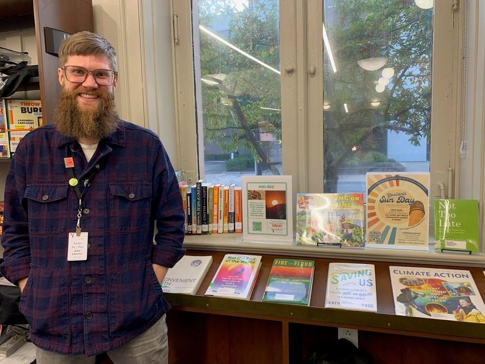 Library staff member Aaron Smith pictured with glasses and plaid shirt standing behind Sun Day display table featuring climate and environmental books including titles on solar power, climate action, and sustainability