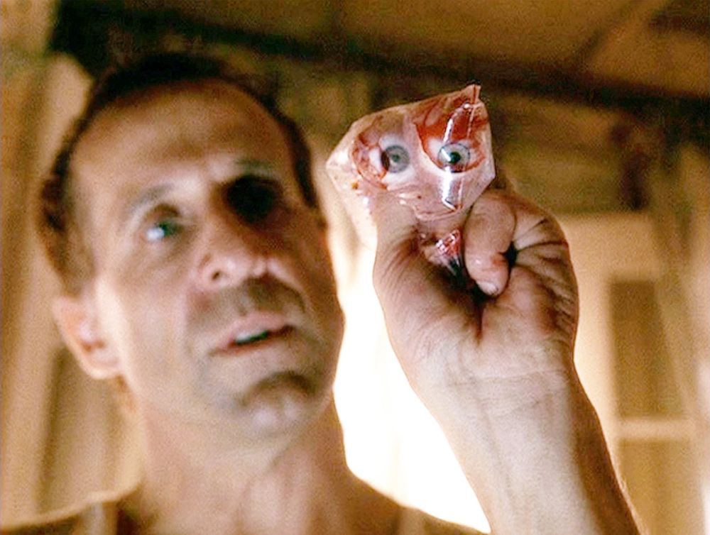 That's a still image from the science fiction film Minority Report. 

The image shows a male, looking directly at the camera, holding a plastic bag containing what appears to be a pair of human eyes and some bloody tissue. His expression is one of shock, disgust, or perhaps contemplation. The background is blurry, suggesting an interior space that is dimly lit. The overall tone is dark and unsettling.