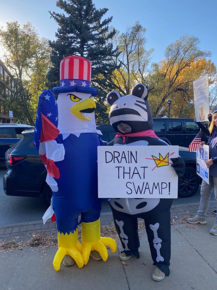 A person in an inflatable skeleton frog holding a sign that says, "DRAIN THAT SWAMP!" stands next to an inflatable red white and blue inflatable eagle.