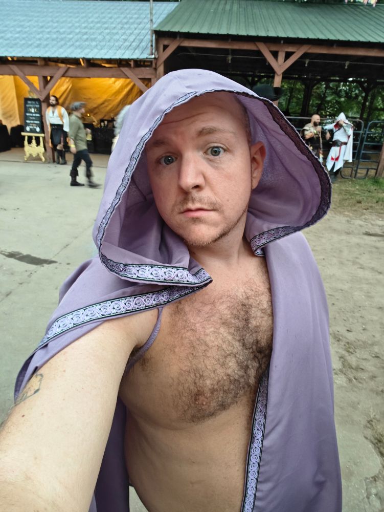 Selfie of me shirtless wearing a purple cloak. There are barn-type areas behind me 