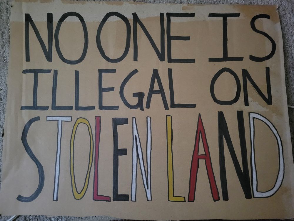 Photo of a cardboard sign that reads "No one is illegal on stolen land." The text is in black except for the final two words, which are written in the alternating colors of the medicine wheel (black, white, yellow, red)