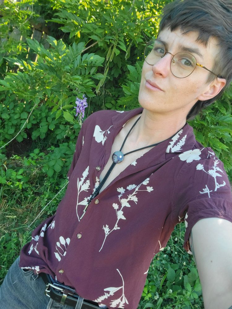 A selfie of a white nonbinary transmasc person with brown hair cut into a mullet style. They are wearing round glasses, a maroon button down shirt with white botanical illustration silhouettes on it. The shirt is unbuttoned at the top and they are wearing a bolo tie 