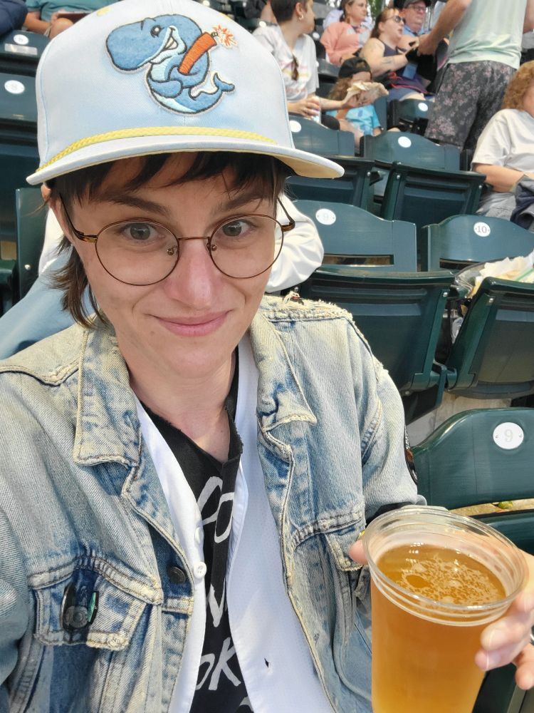 A selfie of a white nonbinary transmasc person with brown hair and round glasses holding a beer and wearing a jean jacket and a light blue baseball hat with an embroidered logo of a cartoon whale holding a stick of dynamite 