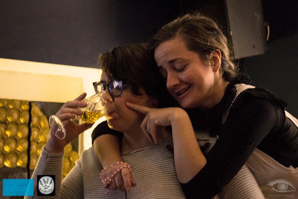 The same two women in a coloured photo. One of the women is drinking from a wine glass the other is behind her one arm wrapped around her neck with the rosary beads around her hand and the other hand is poking her cheek. The woman from behind looks drunk.