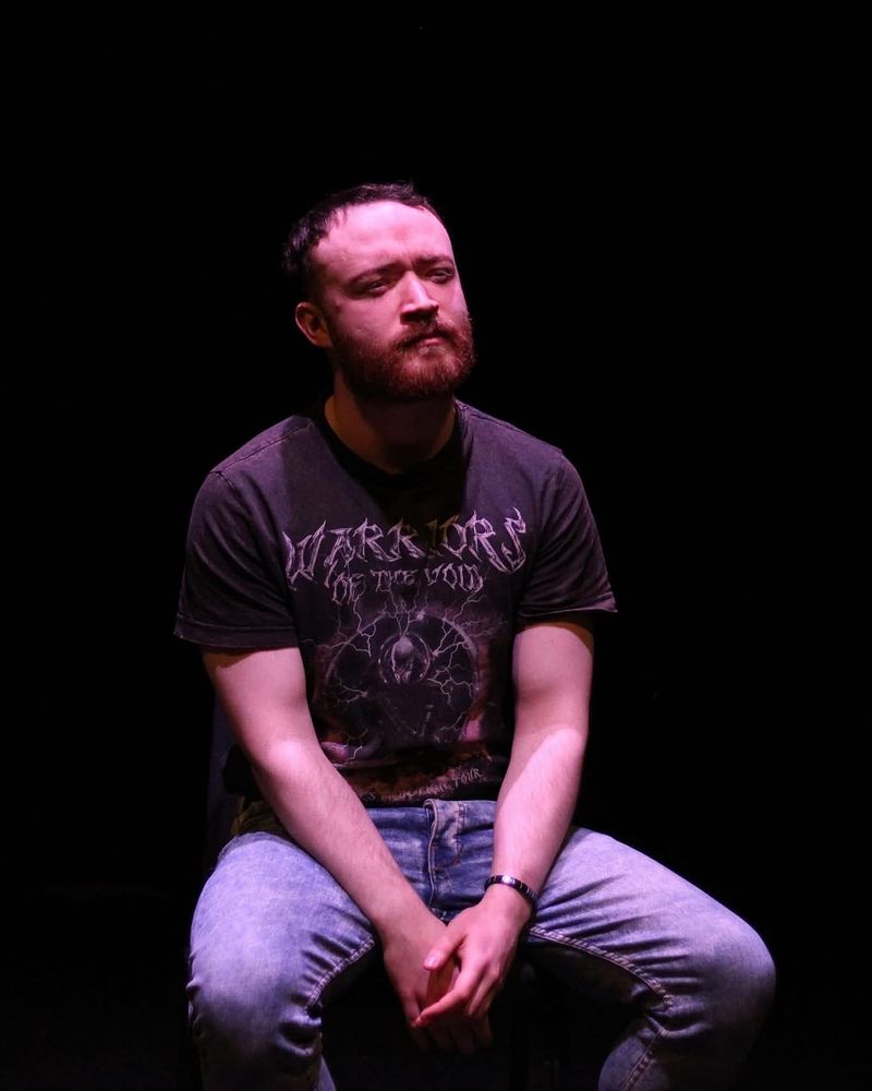 A man is sitting on a stage. He’s wearing jeans and a top. 