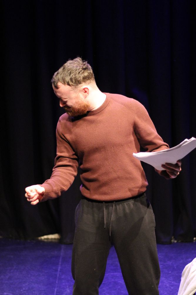 The man standing with a script in one hand and the other hand in a fist. He has his head turned away looking at the side. His face is screwed up.