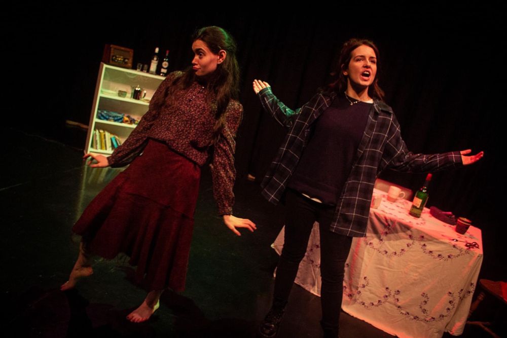 The woman in the red skirt looks off stage.

She is dancing. Beside her, the other woman has her arms displayed talking to the audience.