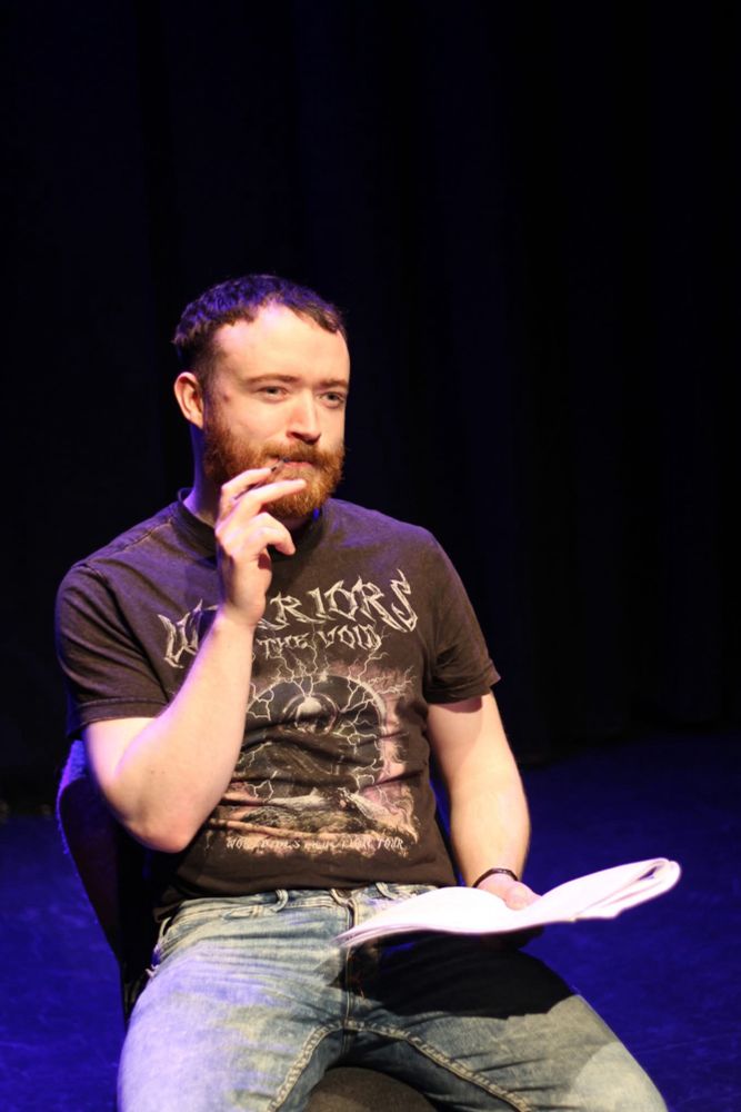 A man sits on the stage, holding a pen to his mouth. 