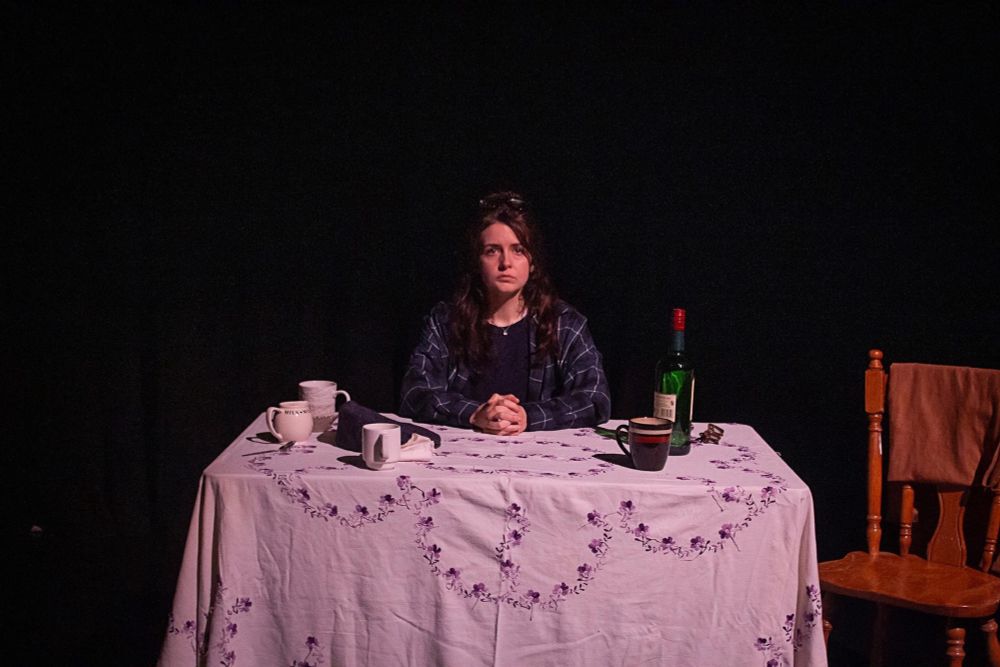 The woman with the open blue shirt is sitting behind a table with a white floral tablecloth upon it. on the table are various cups and a bottle of Jameson. Her hands are classed on the table and she’s looking at the audience. To the right of her is a chair with a brown piece of fabric on it.