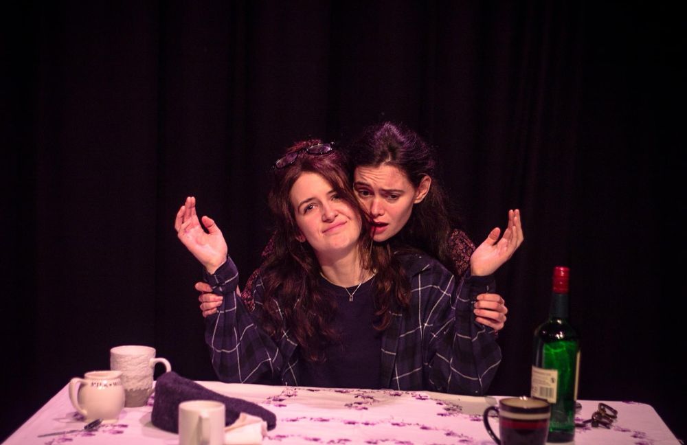 The woman from the fourth sits behind the table she looks at the audience, her face slightly annoyed. Behind her, the other woman is clutching her hands and raising them up slightly.