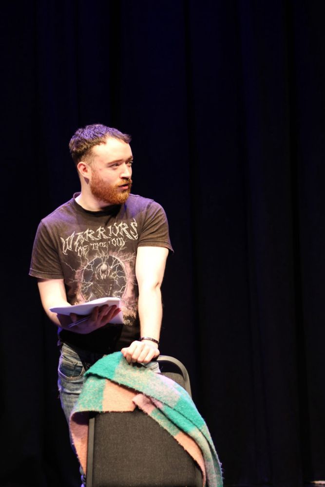 A man stands behind a chair on a black stage. He is looking to the left. 