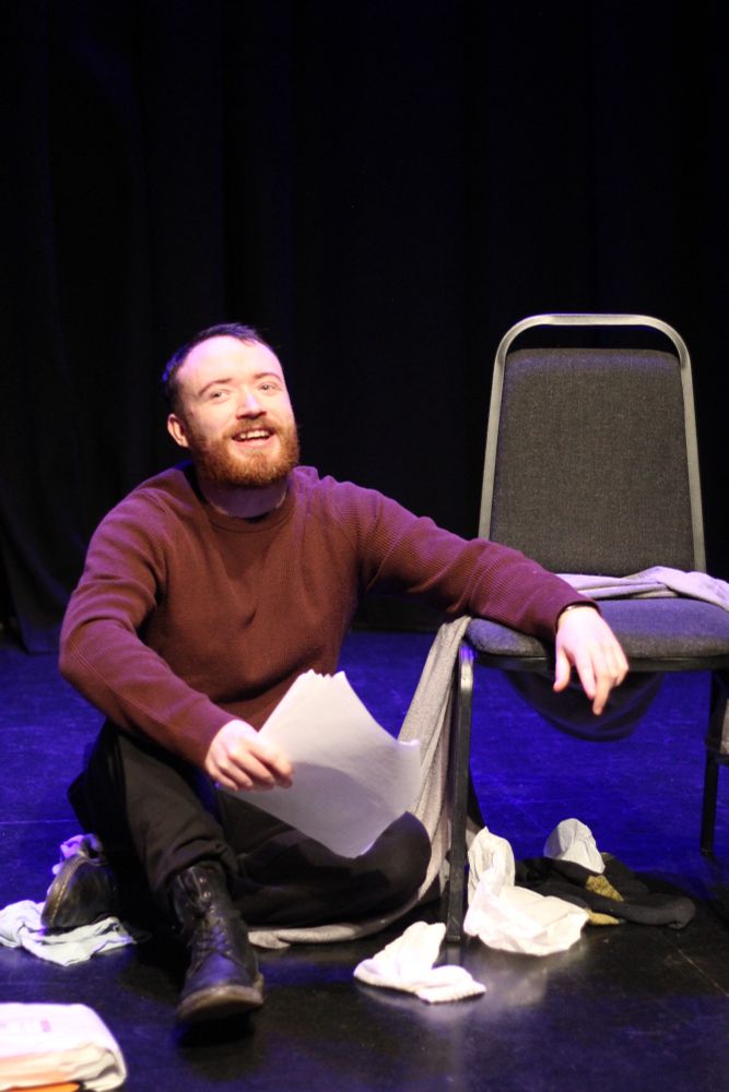 A man sits on the ground and rest his arm on the chair. There are bits of rubbish surrounding him on the black floor and he’s smiling.
