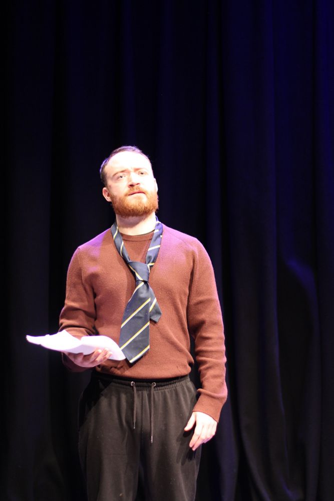 A man stands looking out at the audience. He has a tie wrapped loose around his neck.