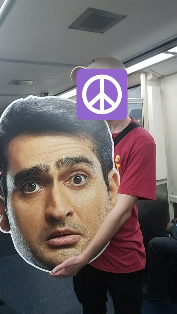 A man in a red t shirt holding a giant cardboard cut out of Kumail Nanijani's head.