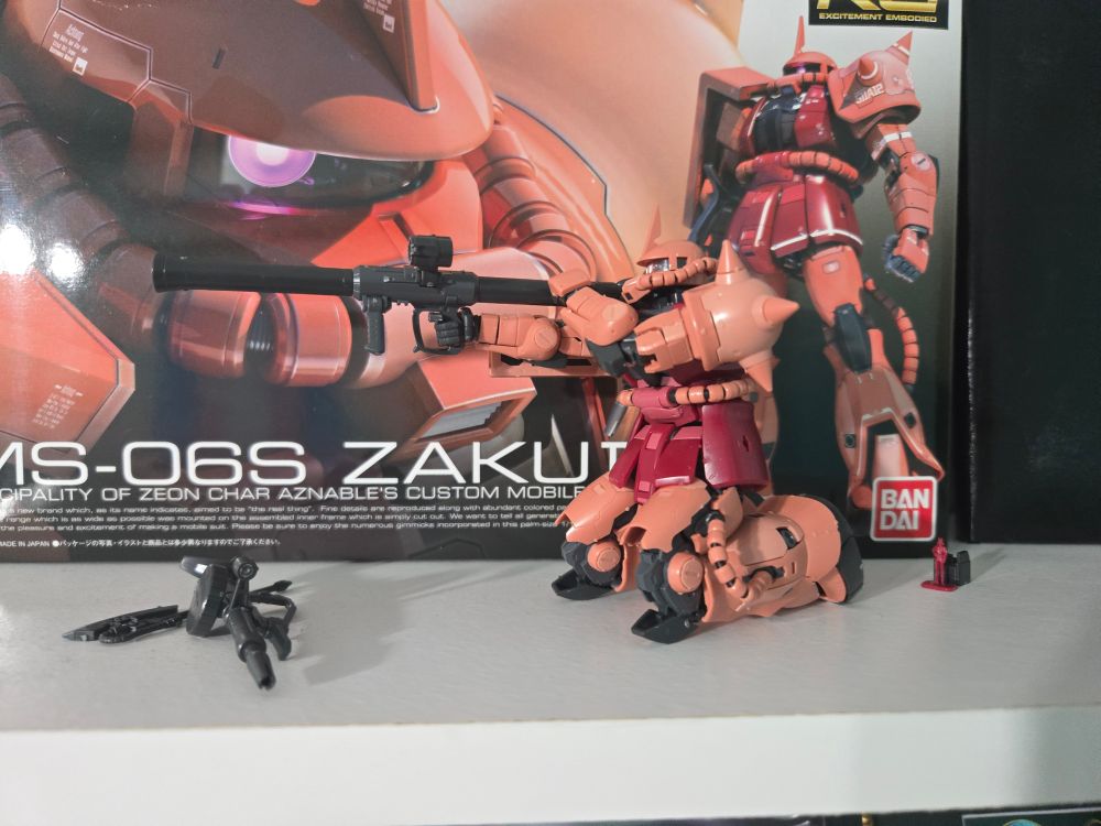 The same one eyed, red mecha with a horn, kneeling down and holding the rockey launcher ready to fire, using its other arm to brace the back of the launcher