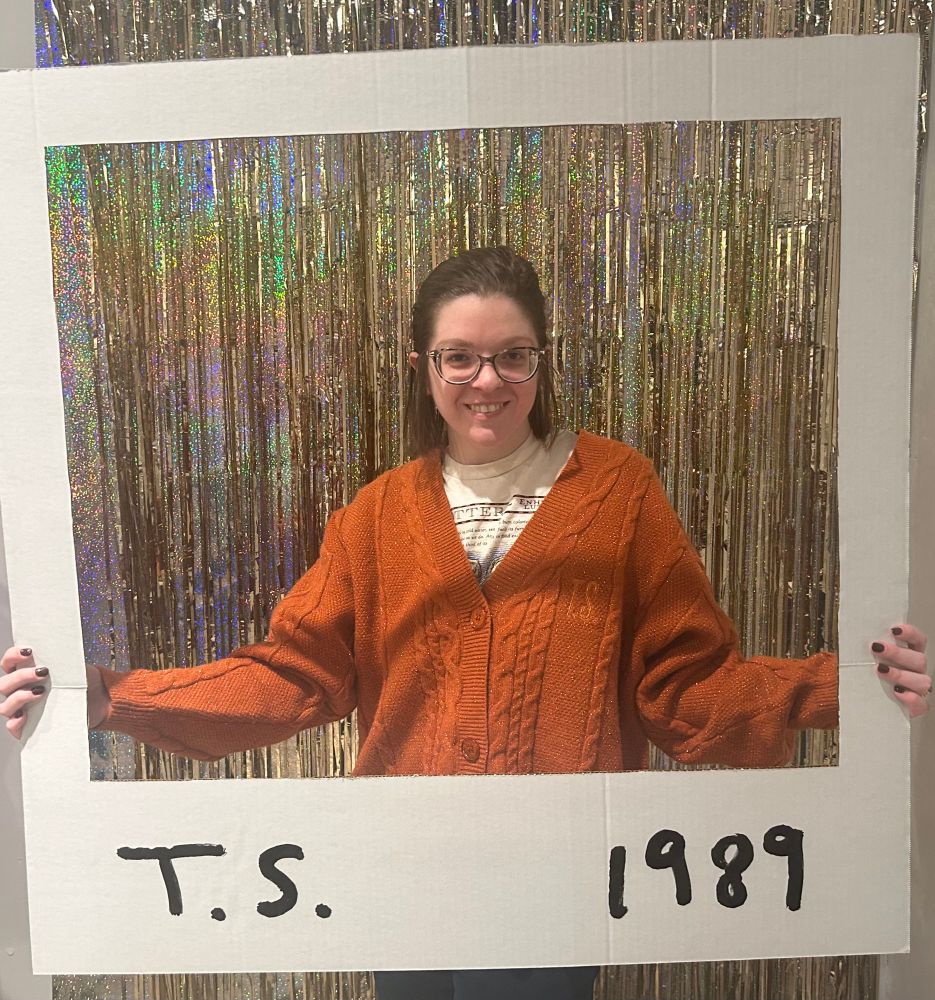I am standing in front of a glittery gold background and holding a giant Polaroid that says T.S. 1989 at the bottom. I am wearing a Monterey bay otter shirt and life of a showgirl orange cardigan